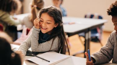  girl laughs happily in a primary school class, she is sitting at a table with her her classmates.