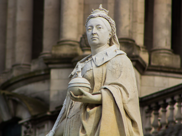 Statue of Queen Victoria positioned in front of a historic stone building.