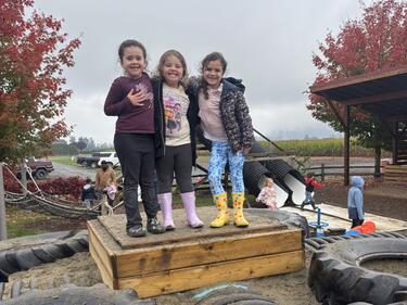 Three young elementary students stand on a wooden box with smiling faces as they pose for a photo at the pumpkin patch.
