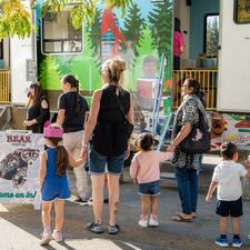 Group of adults holding childrens hands as they wait outside an RV that has been transformed into a play place.